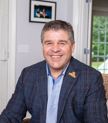 Smiling man in a plaid blazer sits in a chair, with a framed photo on the wall behind him.