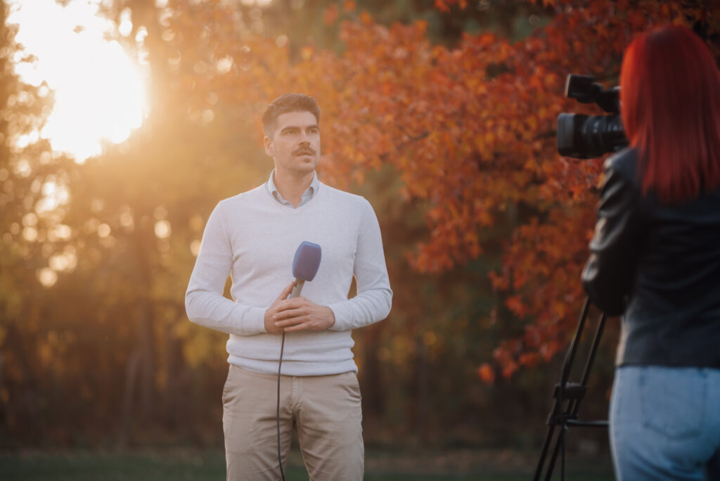 Reporter holding microphone being filmed in autumn park at sunset Male reporter holds a microphone while being filmed by a camerawoman in an autumn park at sunset.