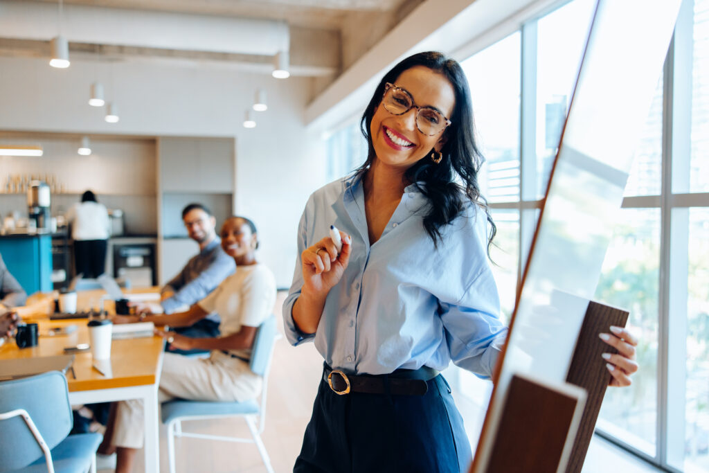 A smiling woman presents to engaged coworkers in a bright office meeting space.
