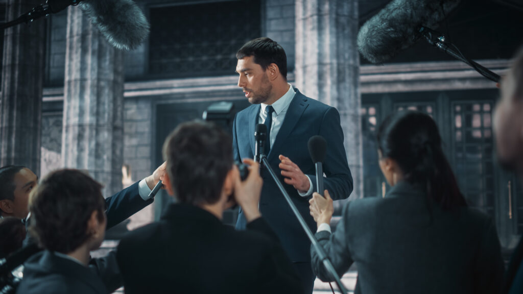 Young Organization Representative Answering Press Questions and Giving Interview Outside a Parliament, Court or Other Government Building. Press Officer or Businessman Crowded by News Journalists. A young representative answers questions from journalists outside a government building, surrounded by microphones and cameras.
