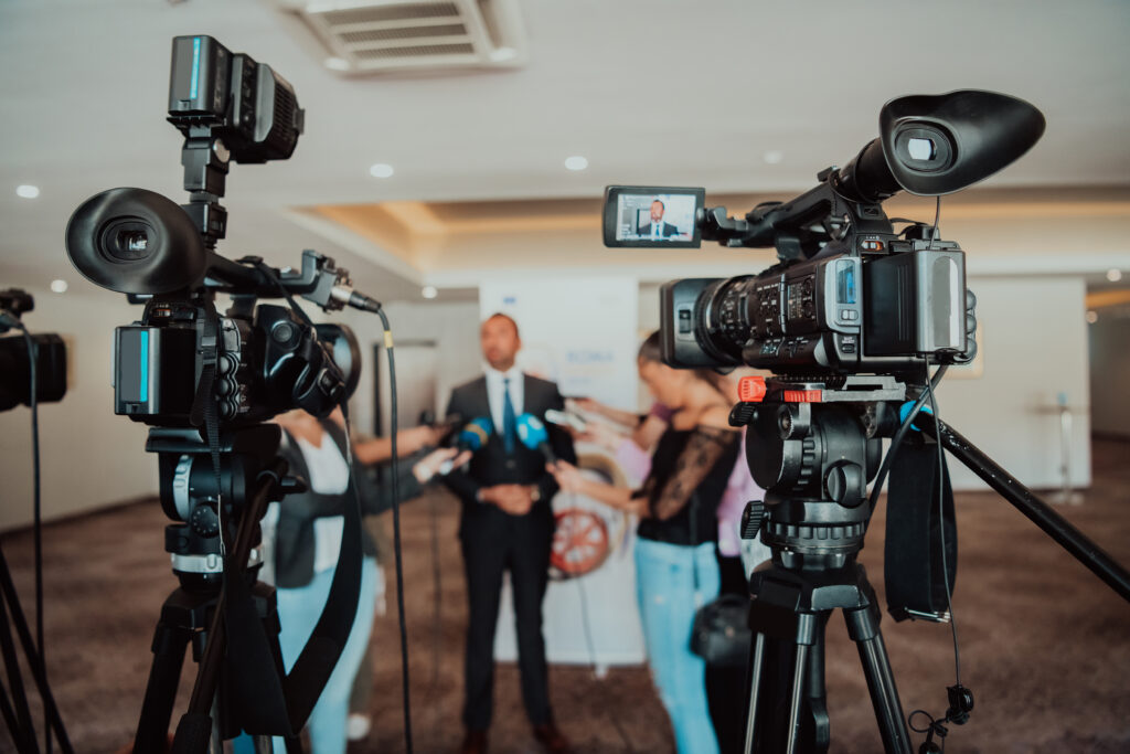 Video cameras capture a speaker surrounded by reporters at a seminar, broadcasting live.