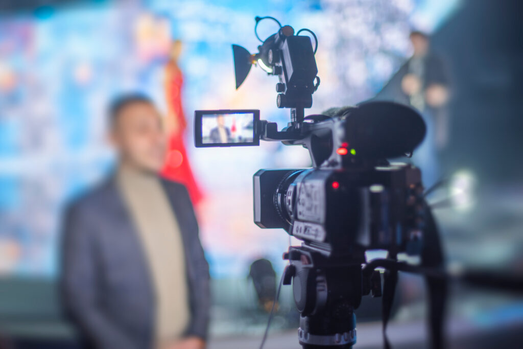 Cameraman films an interview with a blurred subject in the background, focusing on the camera's display.