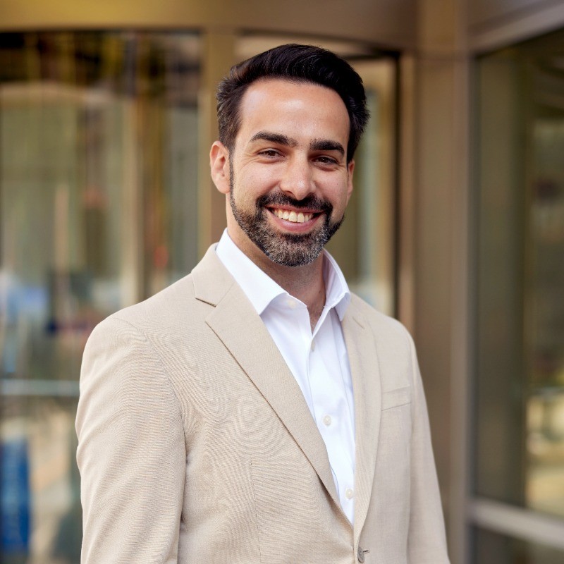 Man in a light beige suit smiles confidently outdoors, with glass doors in the background.
