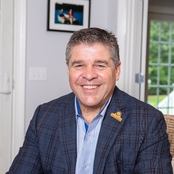A smiling man in a plaid blazer sits indoors, with a framed photo on the wall behind him.