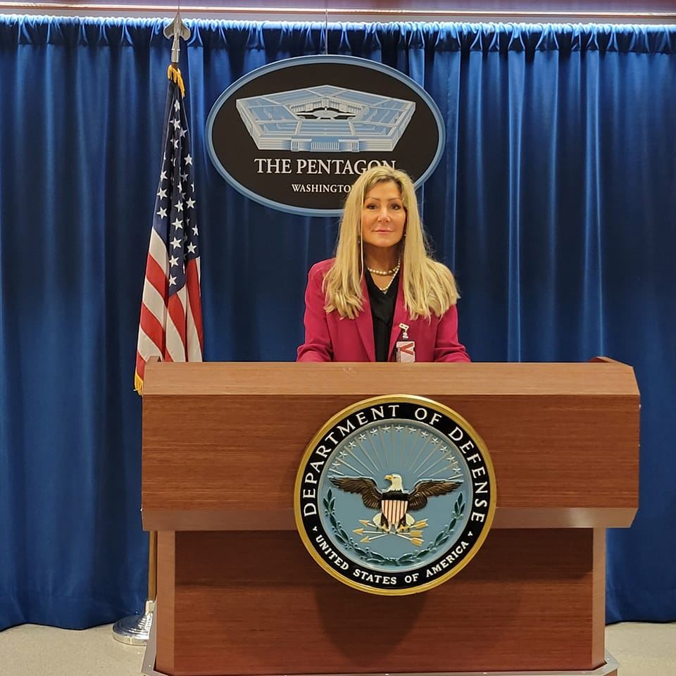 Melissa Travis stands at a podium with the Pentagon seal, U.S. flag in the background.