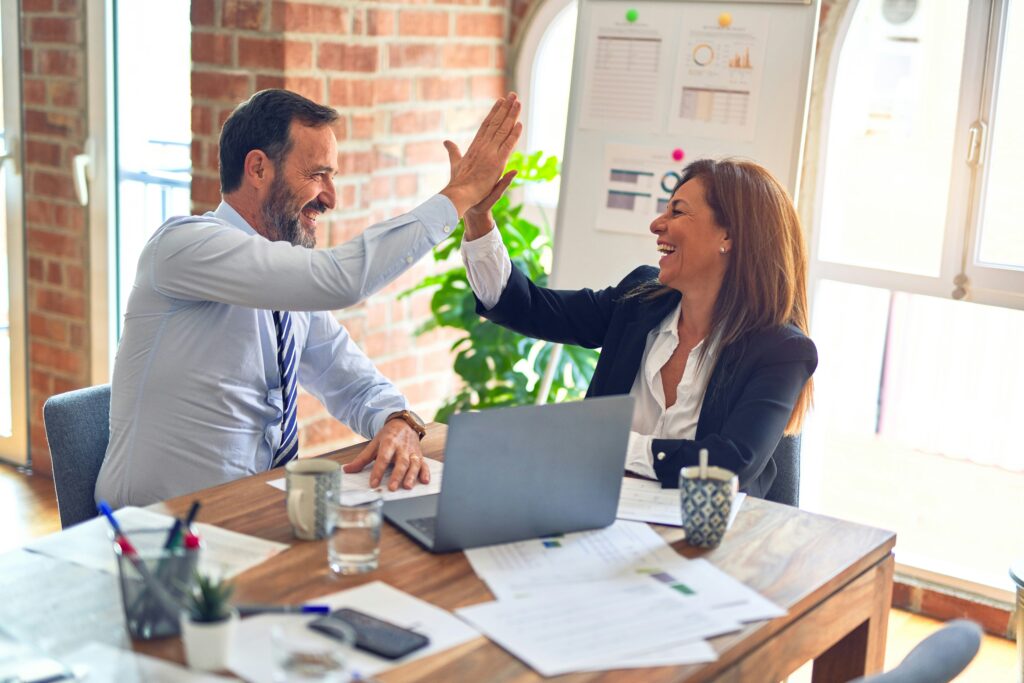 Two colleagues high-five each other at a table with a laptop and documents, smiling in a bright office.